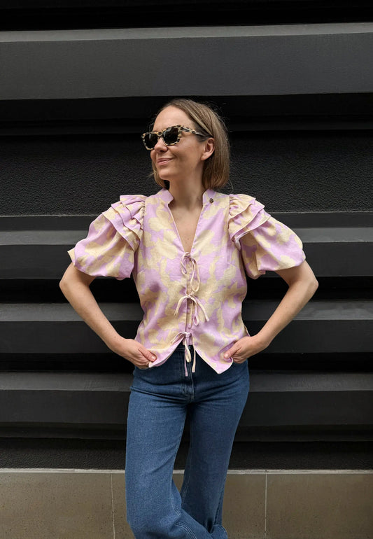 Modelka na tle budynku ubrana w bawełnianą bluzkę ALIANNA wiązaną na przodzie oraz w granatowe jeansy ATLANTA.