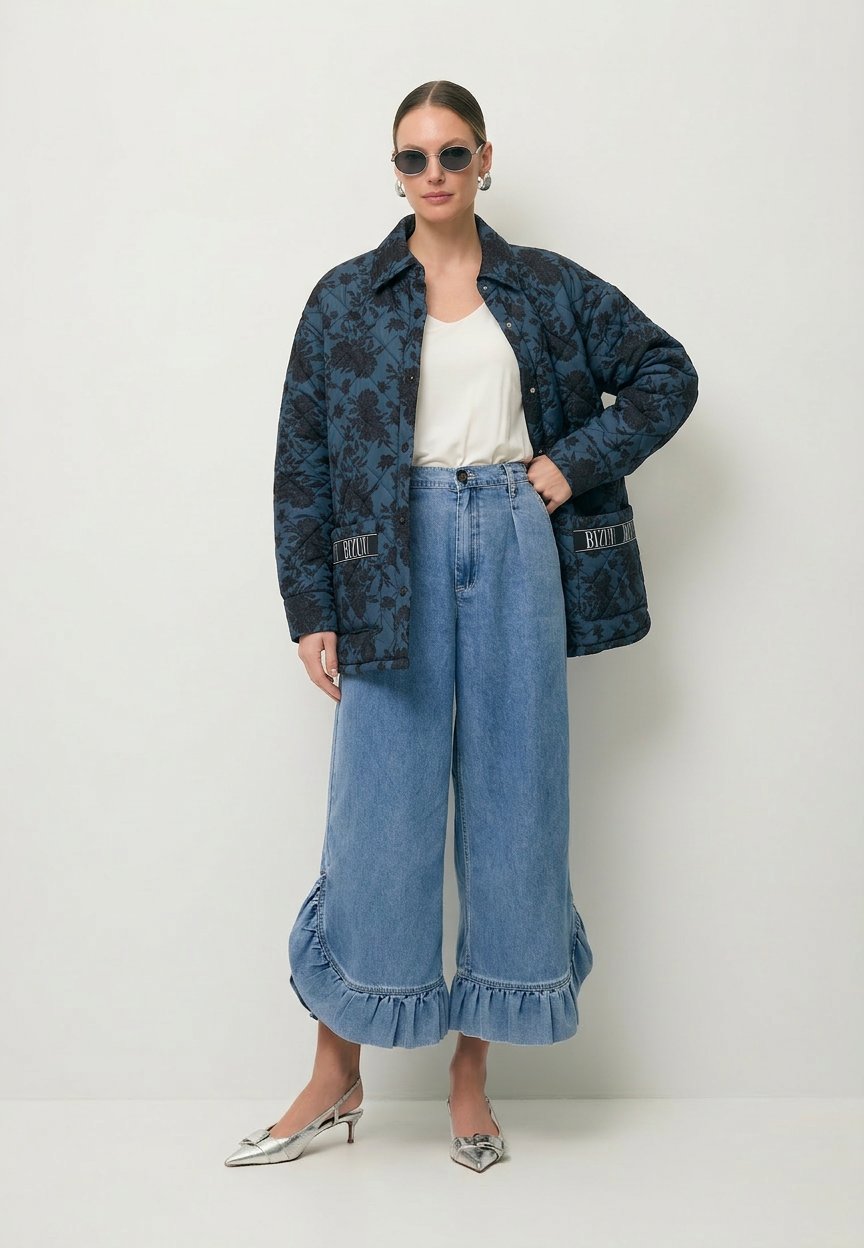 Modelka z ręką na biodrze ubrana w pikowaną kurtkę w print MARIN oraz jeansy z falbaną NELLY.