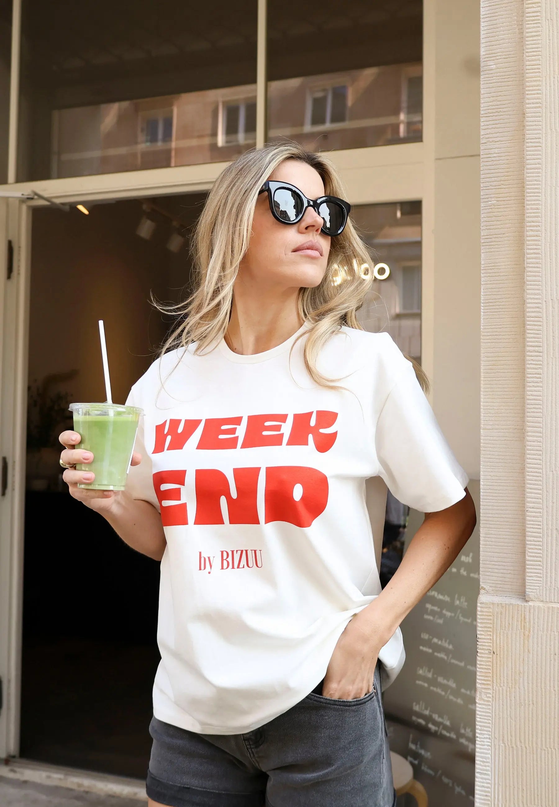 Zbliżenie na modelkę z jedną ręką w kieszeni jeansowych szortów MENORCA trzymająca w drugiej ręce napój. Ubrana w biały T-shirt z czerwonym napisem WEEKEND by Bizuu RALZA.