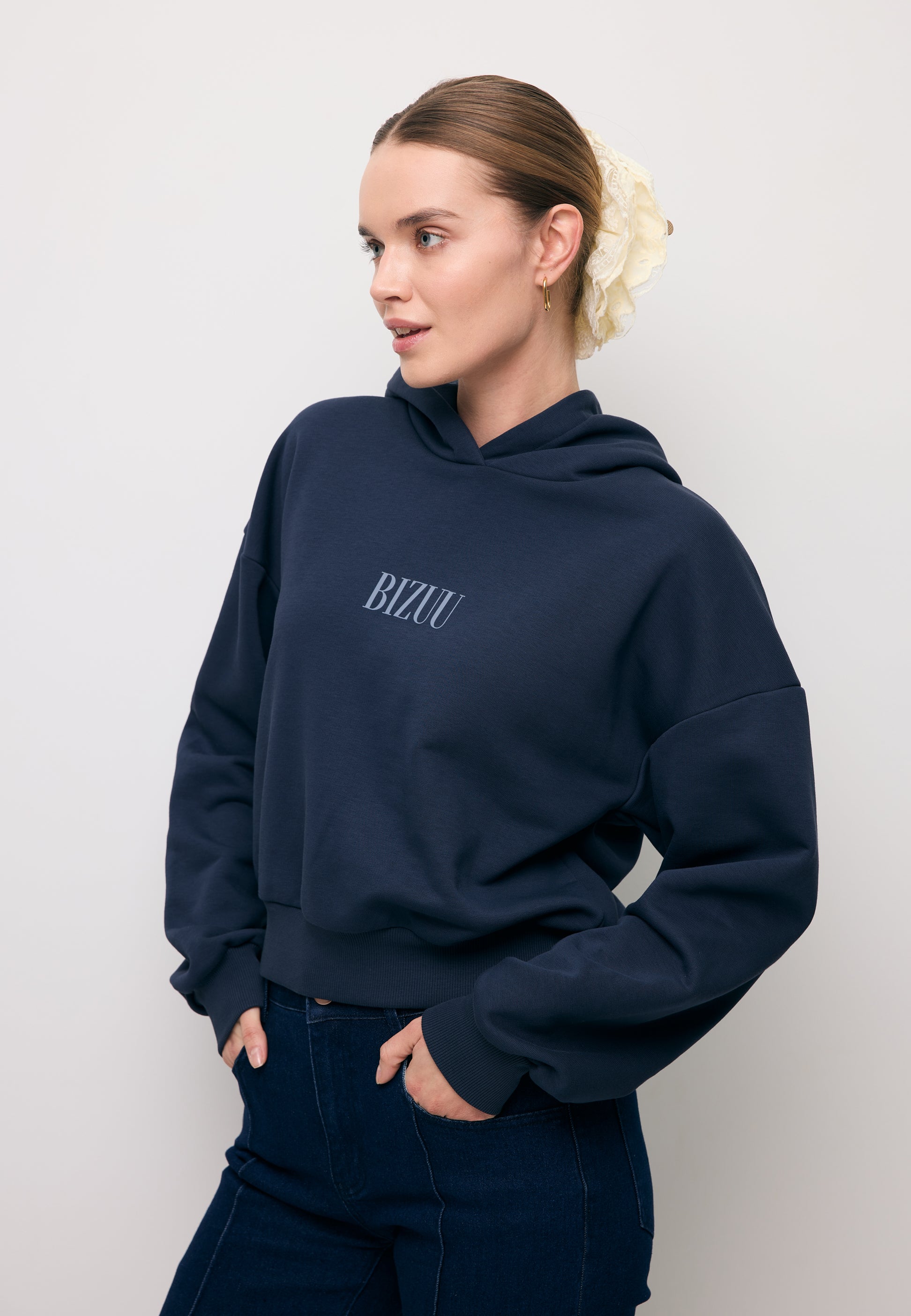 Zbliżenie na modelkę stojącą bokiem z ręką w kieszeni spodni. Ubrana w granatową bluzę z niebieskim logo SAY.