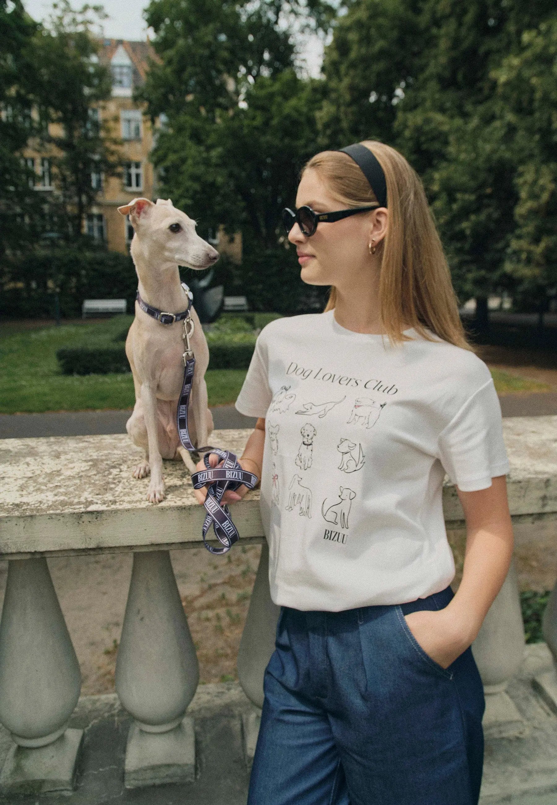 Modelka oparta o murek, na którym siedzi pies. Ubrana w biały T-shirt z rysunkami psów i napisem Dog Lovers Club. W ręce trzyma smycz w logo Bizuu.