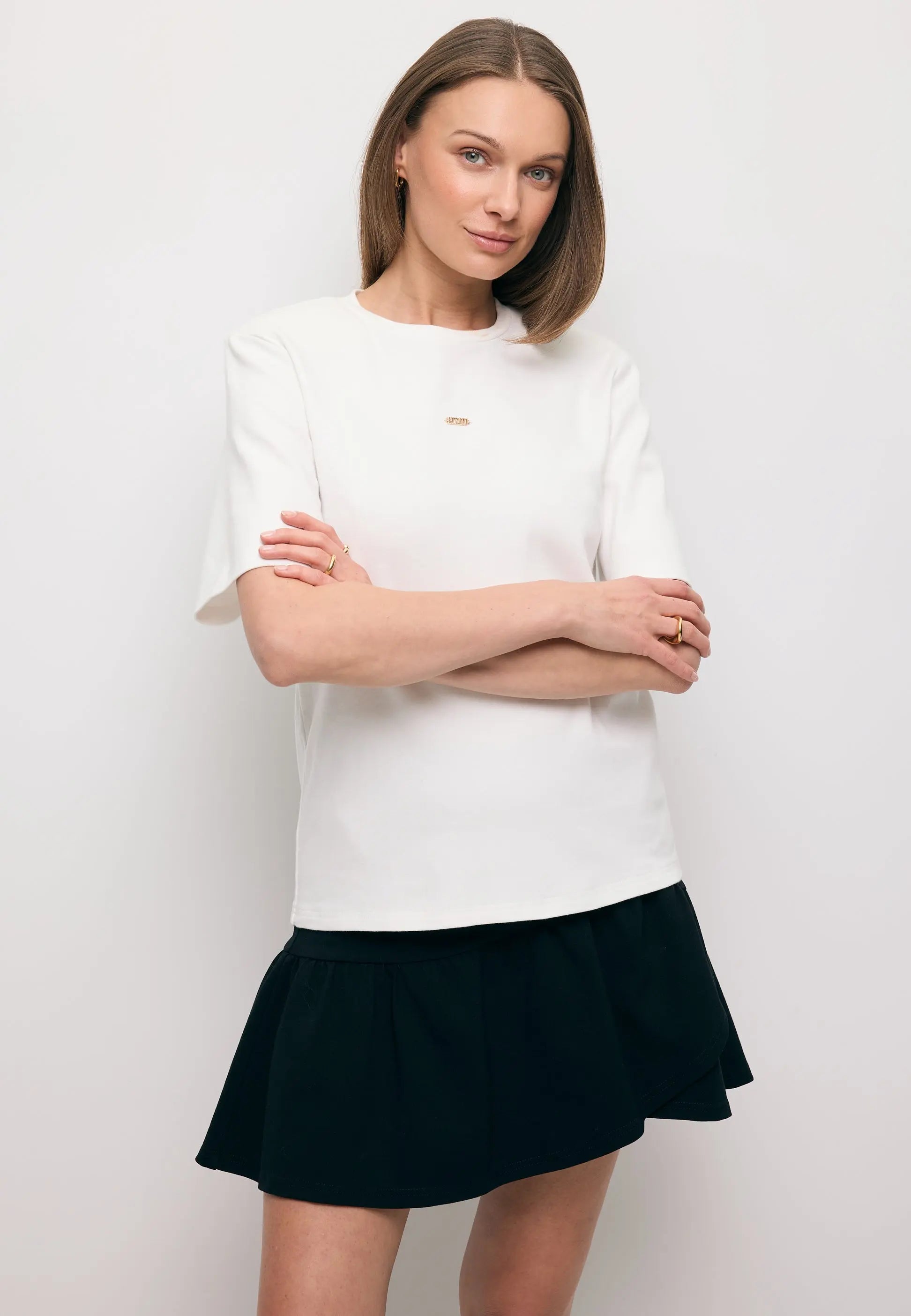 Modelka ze skrzyżowanymi rękoma ubrana w biały T-shirt z ozdobnymi poduszkami MELISA oraz czarną mini spódniczkę TULIAN.
