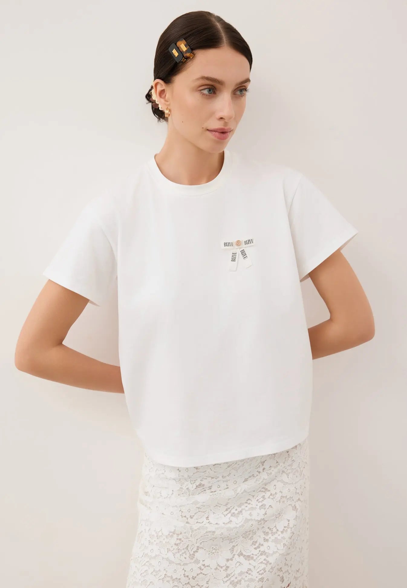 Modelka oparta o białą ścianę z rękoma z tyłu. Ubrana w biały T-shirt z logowaną kokardką TISHALLE i białą, koronkową spódnicę MILKY.
